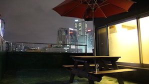 Une terrasse sur le toit, avec une table de pique-nique et un parasol rouge, d’où l’on peut admirer la vue sur la ville illuminée la nuit.