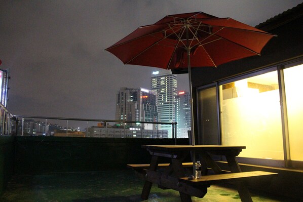A rooftop terrace with a picnic table and a red umbrella, overlooking a cityscape at night.