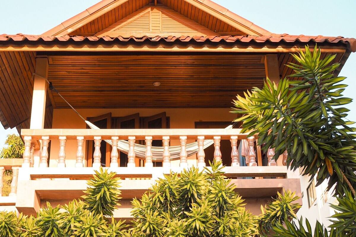 A wooden house with a tiled roof, a balcony with a hammock, and lush green plants around it.