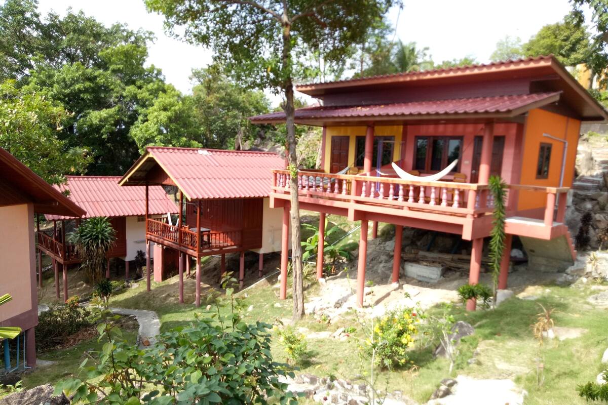 A bungalow with a red roof, a balcony, and a garden area with rocks and plants.