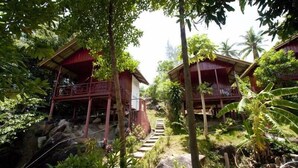Una casa de madera tradicional con balcón, rodeada de abundante vegetación y una escalera de piedra que lleva a ella.