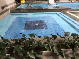 Piscina com duas pessoas na água, rodeada por plantas e um chão de azulejos.