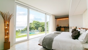 A modern bedroom with a large sliding glass door leading to a pool and garden, a desk with a chair, and a bed with multiple pillows. - Jusandi (Ishigaki)