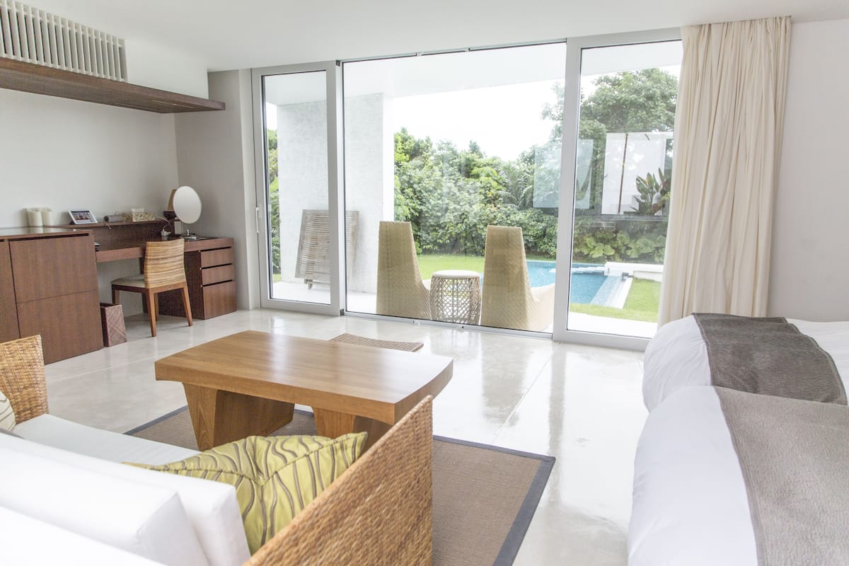 A modern hotel room with a large sliding glass door leading to a pool area, a wooden desk, a sofa, and a dining table.
