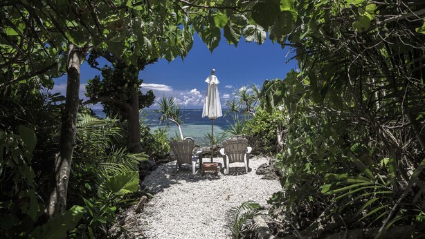 An outdoor seating area with two chairs and a table, surrounded by lush greenery and overlooking a clear blue ocean. - Jusandi (Ishigaki)