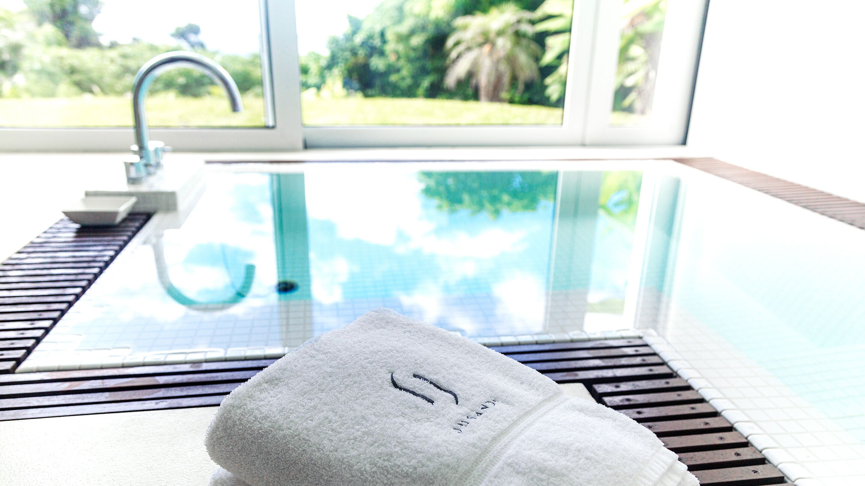 A white towel with a logo on a wooden surface near a glass-enclosed pool with a view of greenery.
