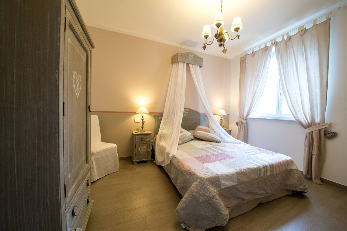 Een slaapkamer met een hemelbed, nachtlampjes, een kledingkast en een raam met gordijnen.