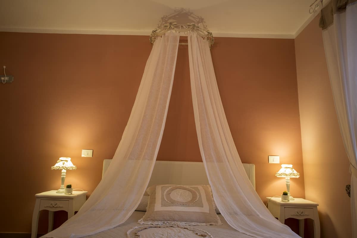 Een slaapkamer met een hemelbed, twee nachtkastjes met lampen en een lichtschakelaar aan de muur.
