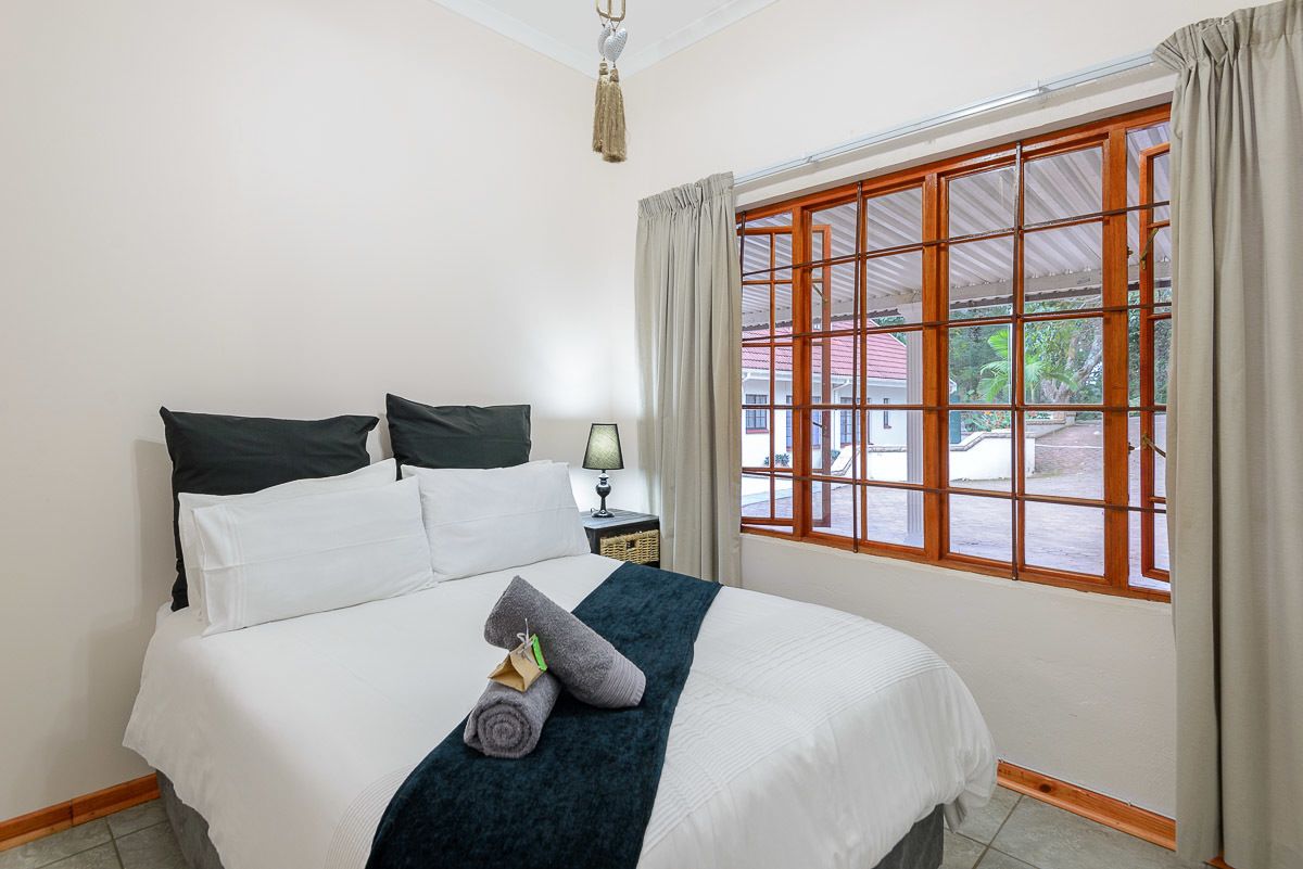 Uma cama bem arrumada com lençóis brancos e verde escuro, uma pequena mesa de cabeceira com abajur e uma janela com cortinas.