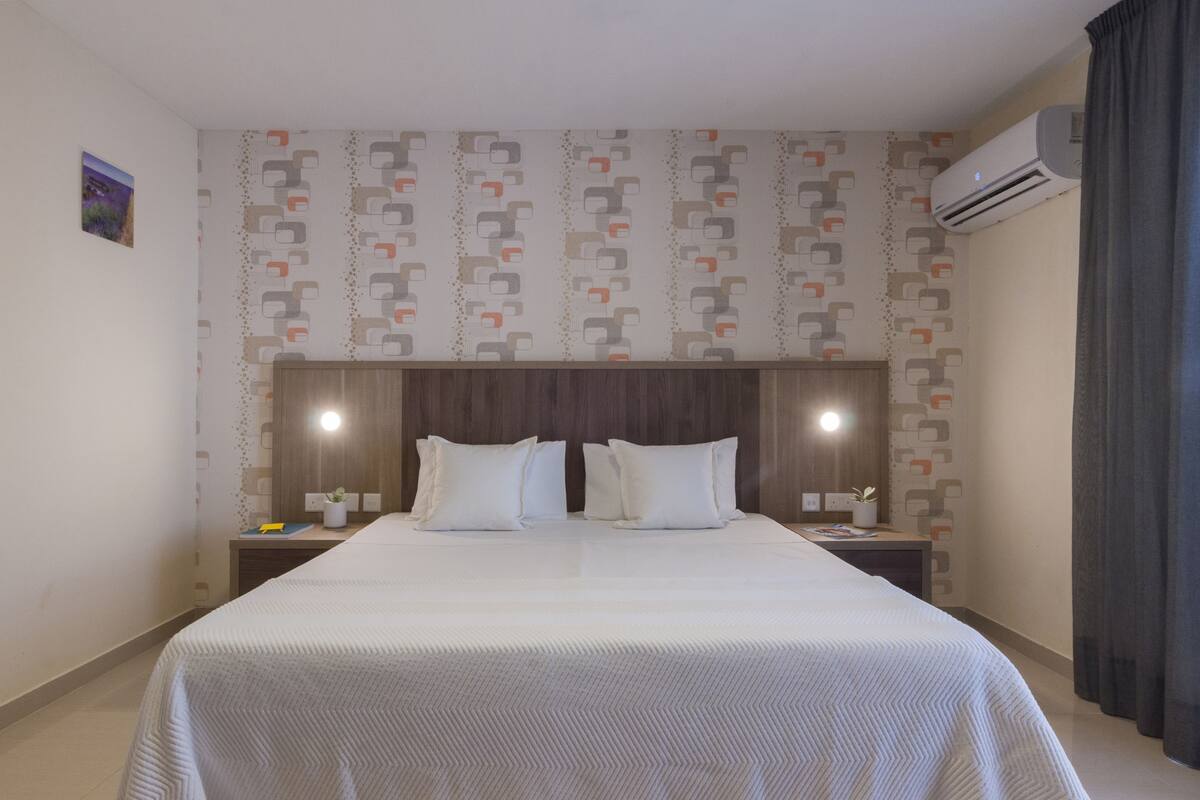 Une chambre d’hôtel avec un grand lit, deux tables de chevet, un climatiseur fixé au mur et un papier peint à motifs.