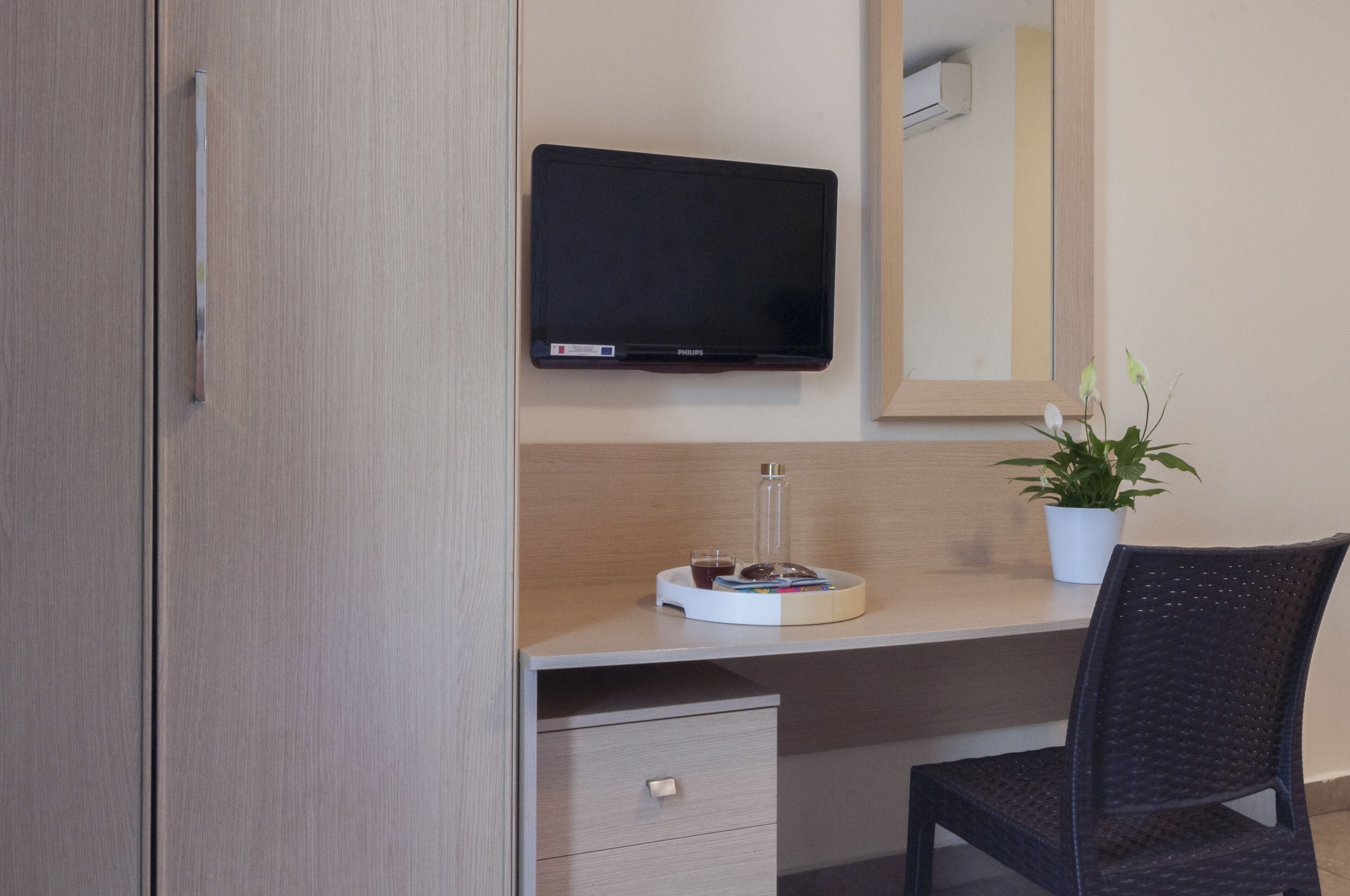 A hotel room with a wooden wardrobe, a desk with a flat-screen TV, a small potted plant, and a chair.