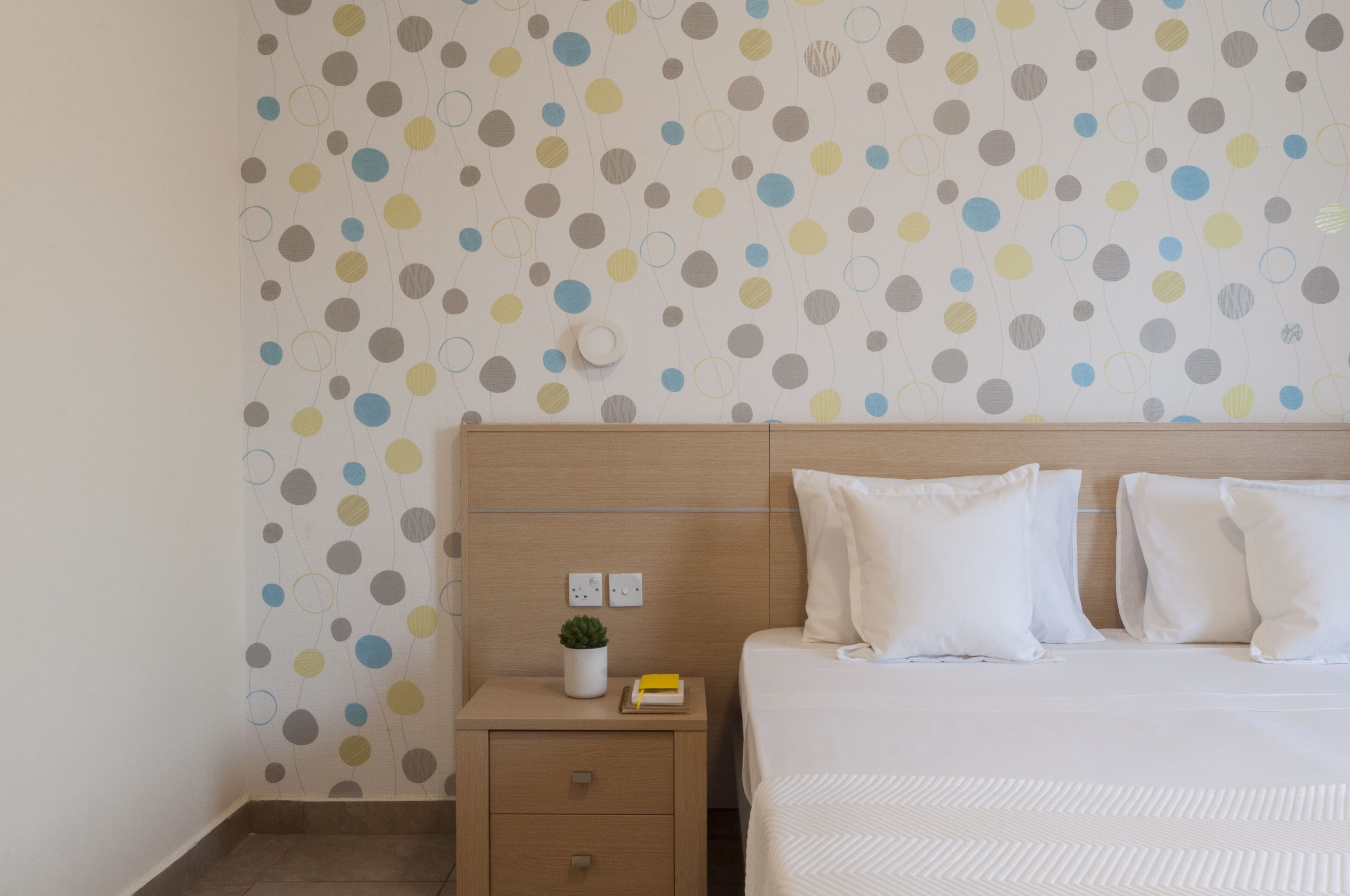 Habitación de hotel con papel tapiz de lunares, cabecera de madera, cama con ropa blanca, mesita de noche con una planta y un libro, y un cajón pequeño.