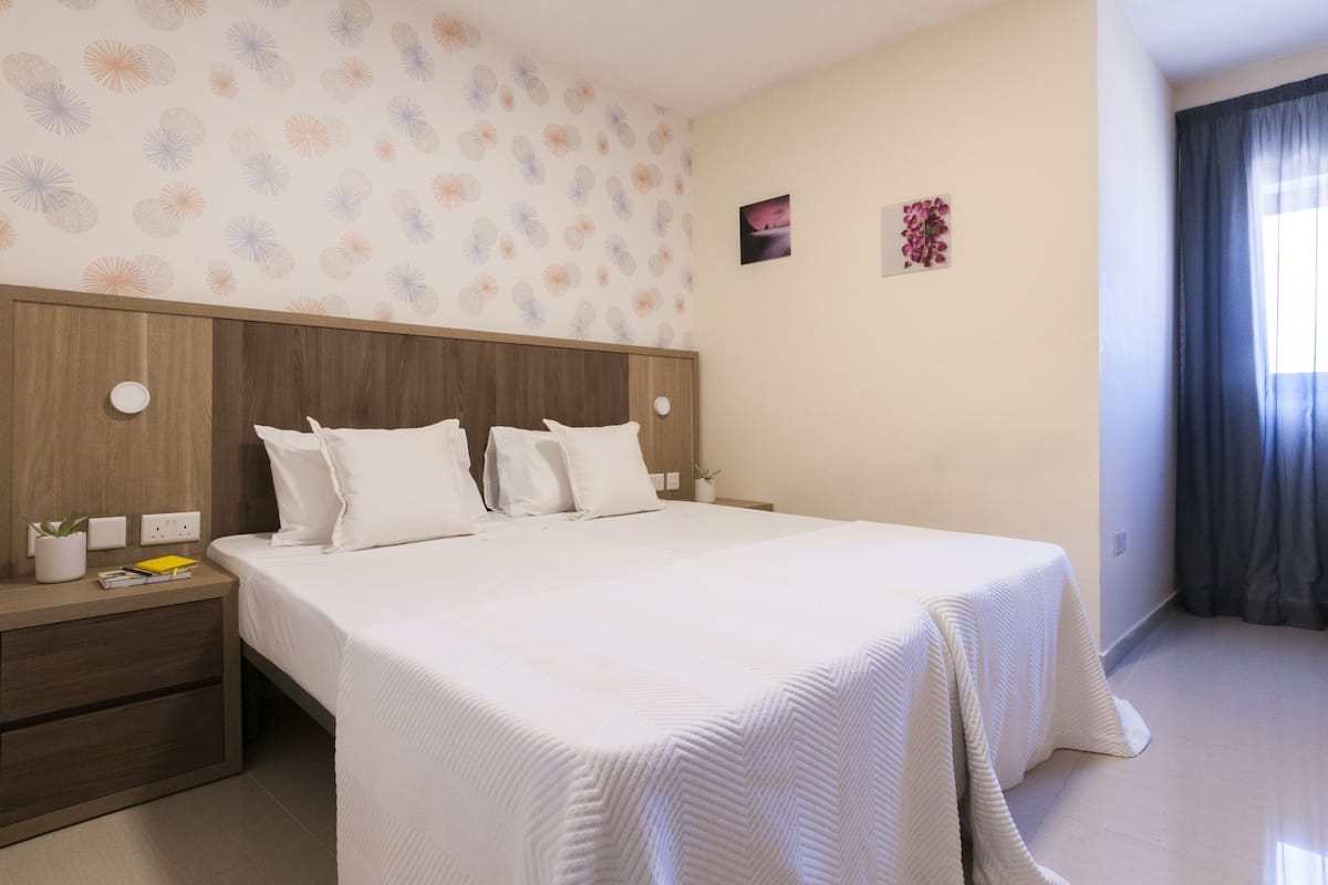 Une chambre d’hôtel avec un grand lit, une tête de lit en bois, une table de chevet agrémentée d’une plante et un mur orné d’un motif floral.