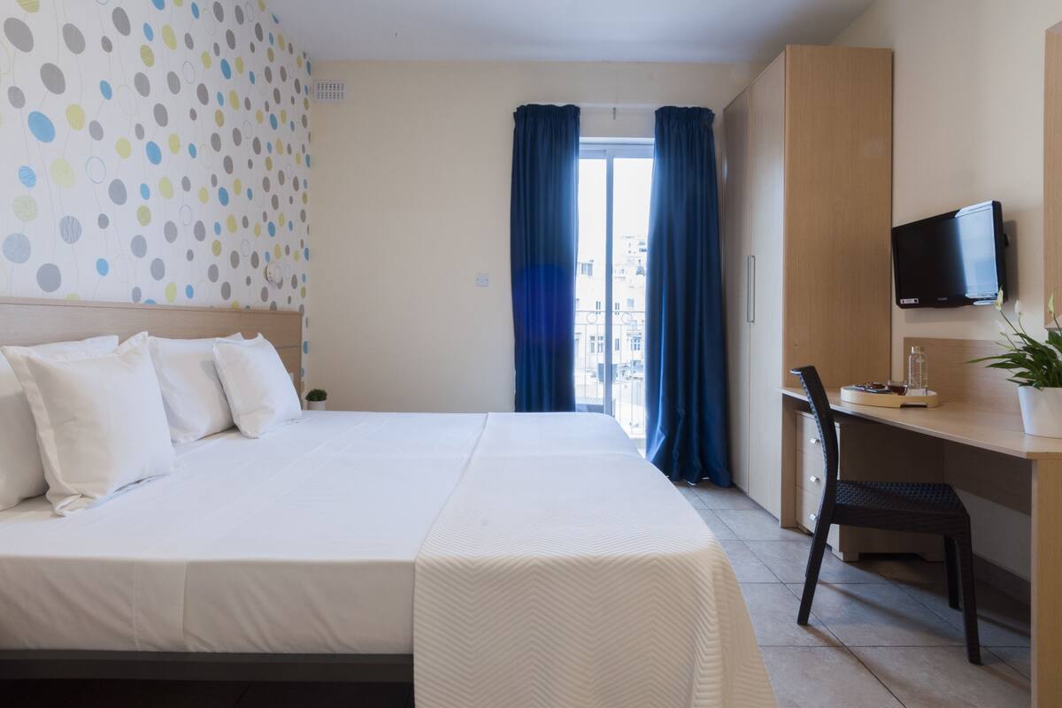 Une chambre d’hôtel équipée d’un lit, d’un bureau, d’une chaise, d’une télévision et offrant une vue sur la ville par la fenêtre.