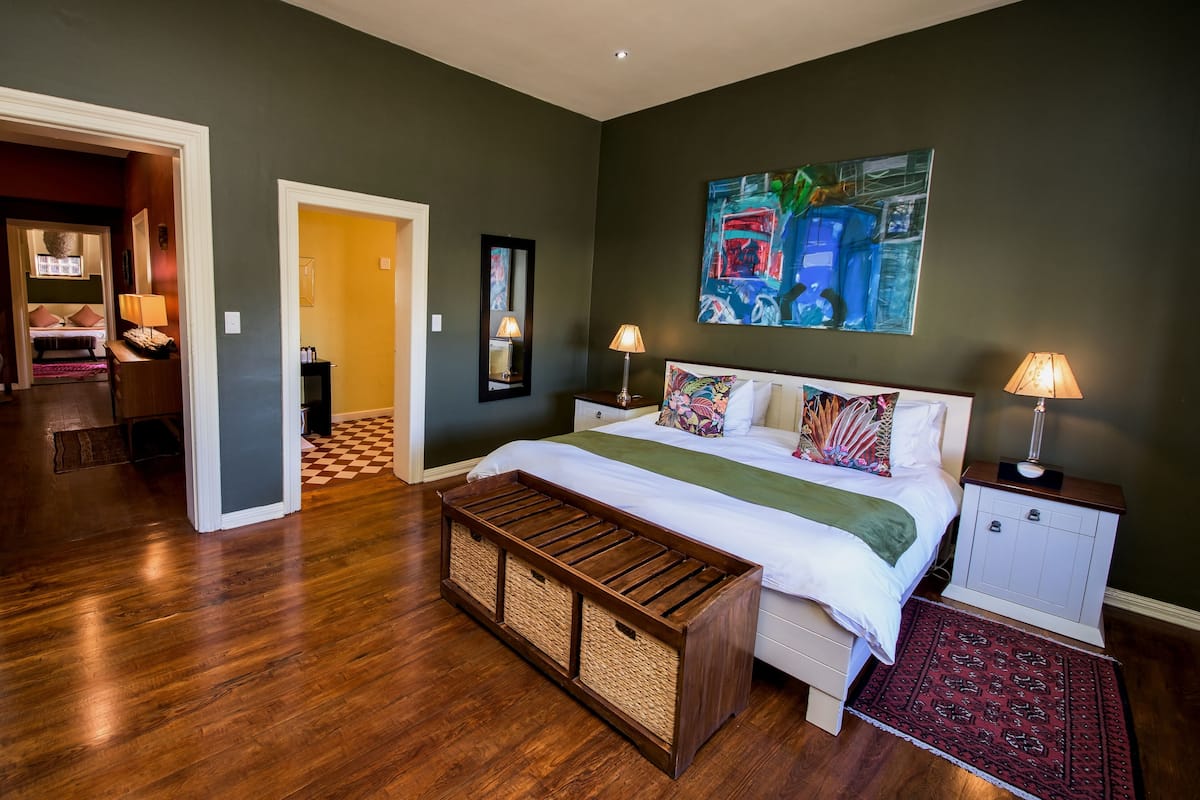 Un dormitorio con una cama, mesitas de noche, un banco, una alfombra, un cuadro y una puerta que da a otra habitación.