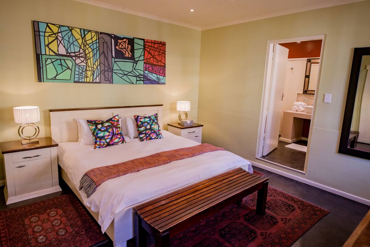 Un dormitorio ordenado con una cama grande, dos mesitas de noche con lámparas, un cuadro abstracto de colores y un baño visible a través de una puerta abierta.