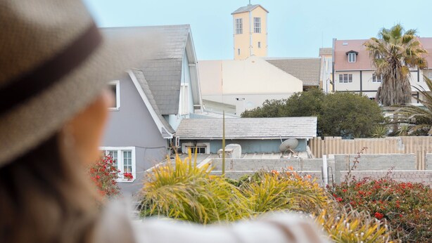 A person in a hat overlooking a residential area with a church tower, palm trees, and a patterned patio. - Villa Margherita (Swakopmund)