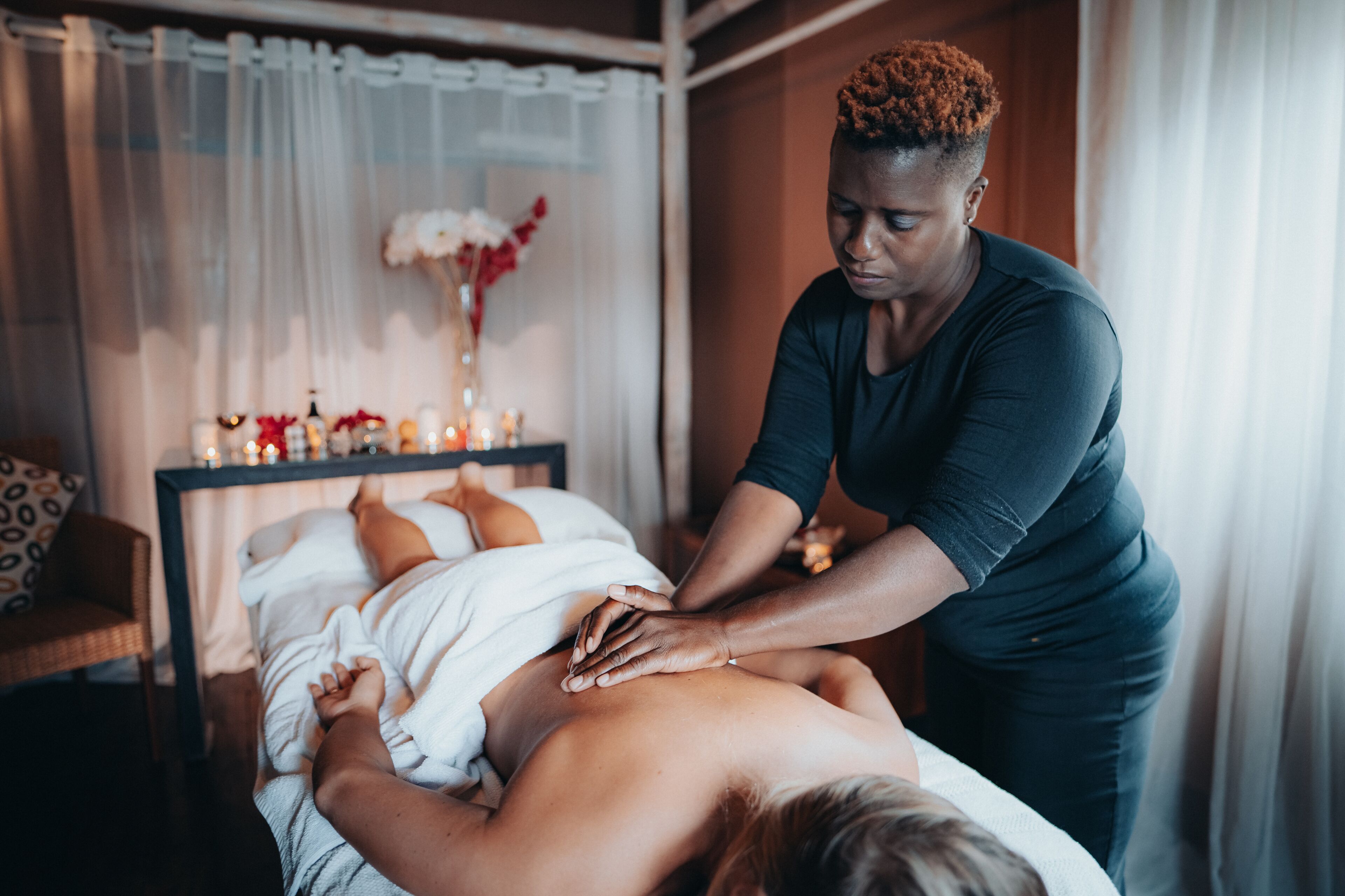 Sessão de massagem em um quarto com uma cama, uma mesinha com velas e um vaso de flores.