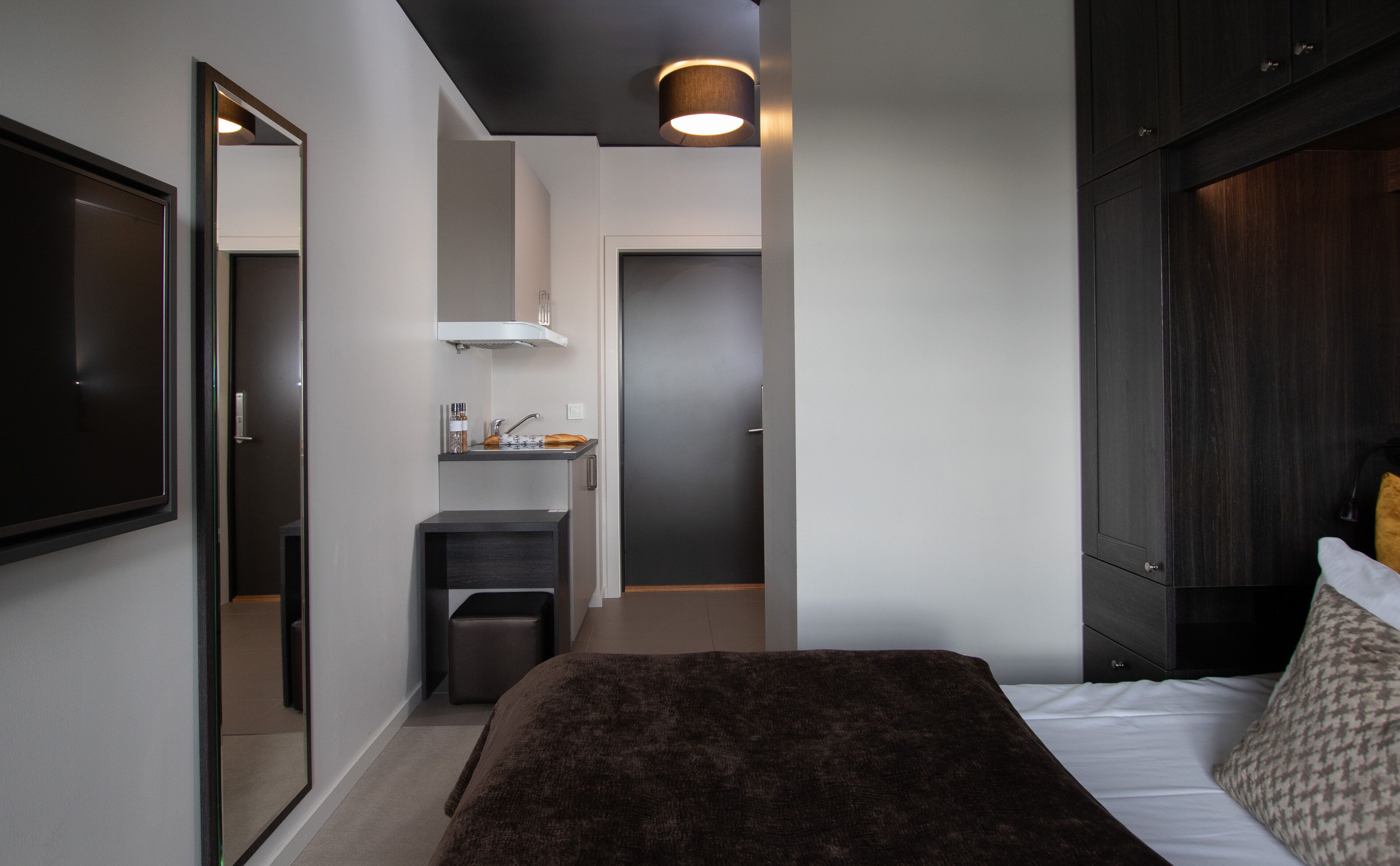 Un dormitorio moderno con una cama grande, cabecera oscura, una pequeña kitchenette y un espejo.