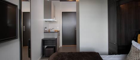 A modern bedroom with a large bed, a dark headboard, a small kitchenette, and a mirror.