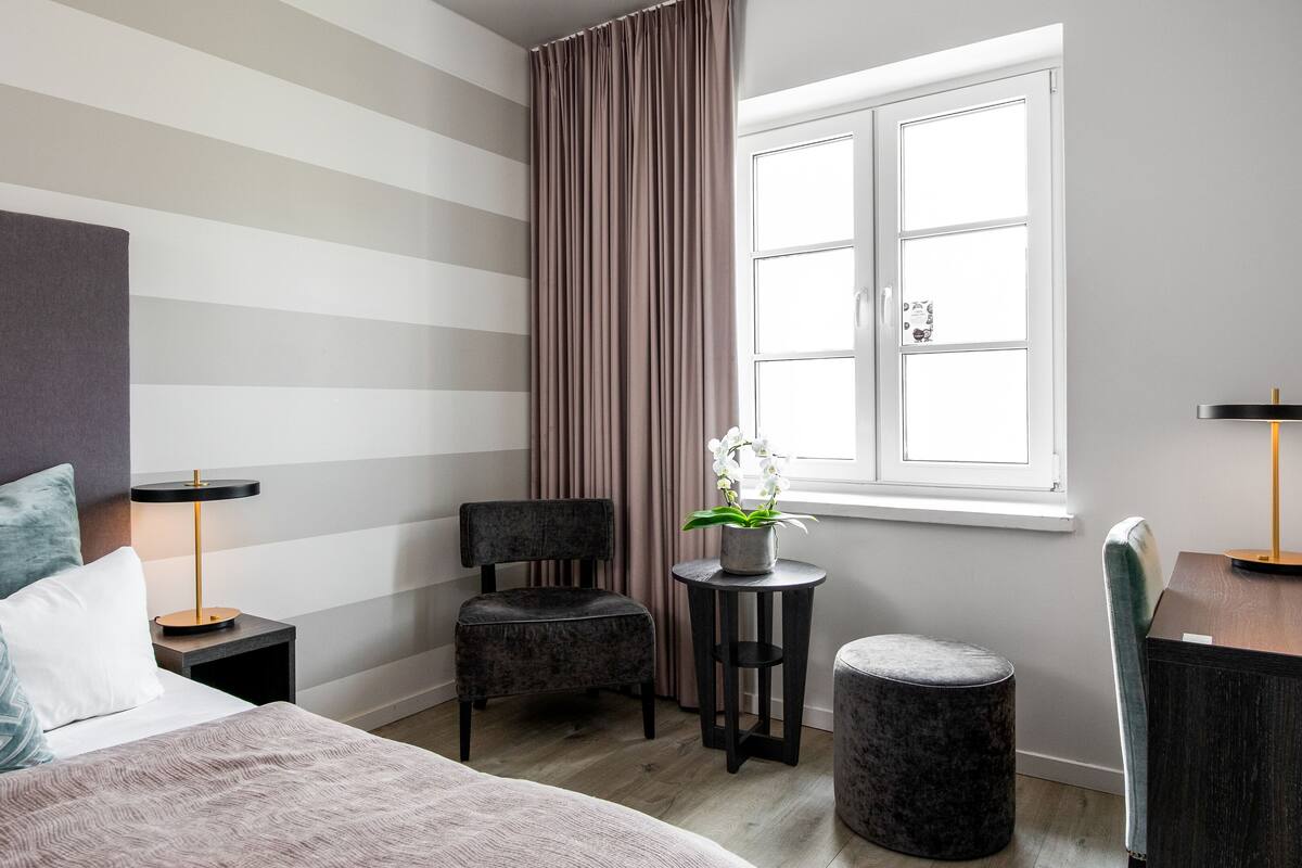 Ein modernes Hotelzimmer mit einem Bett, einem Sessel, einem kleinen Tisch, einem Schreibtisch, einer Topfpflanze und einem Fenster mit Vorhängen.