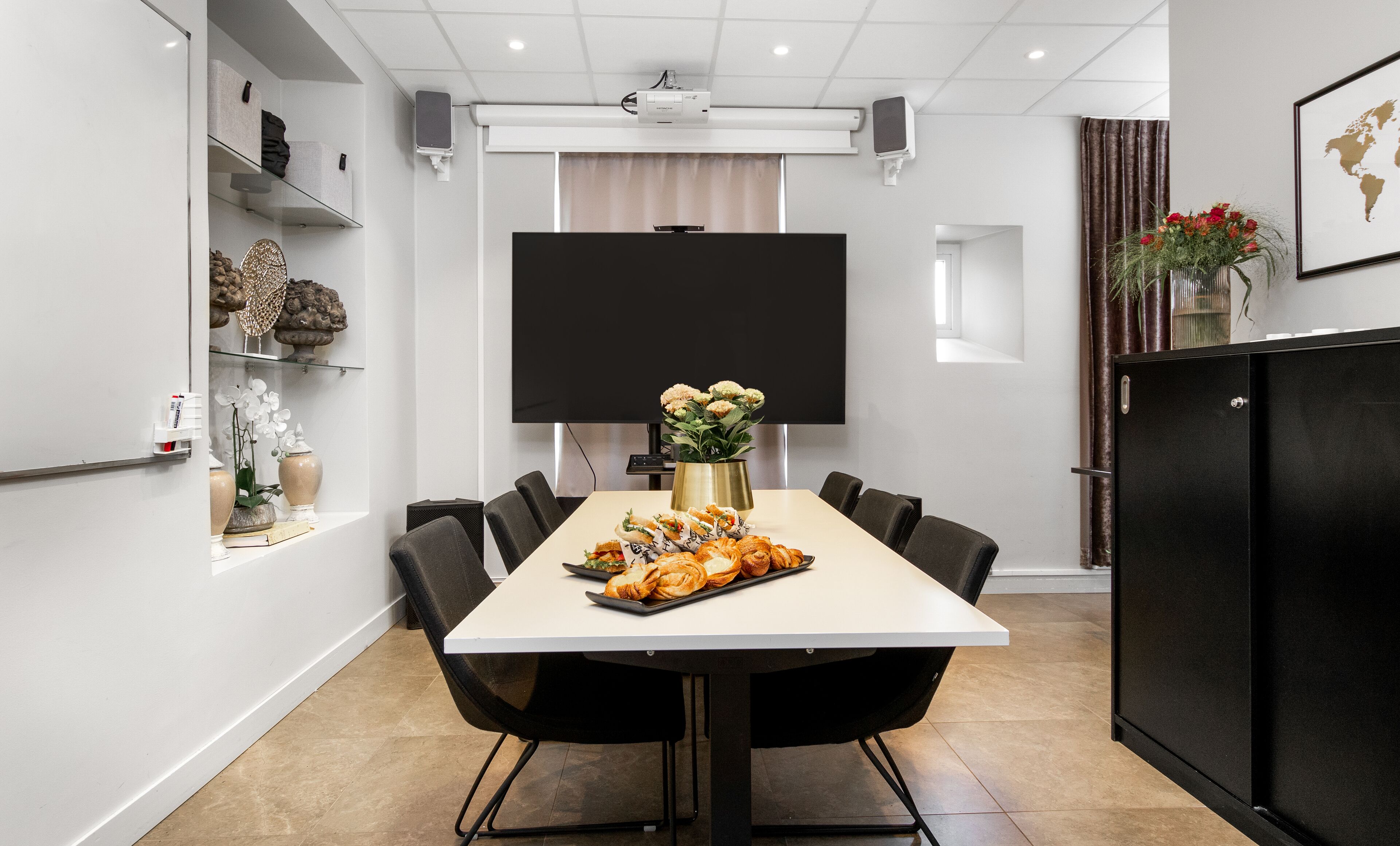 A conference room with a white table, black chairs, a projector screen, and a flower vase.