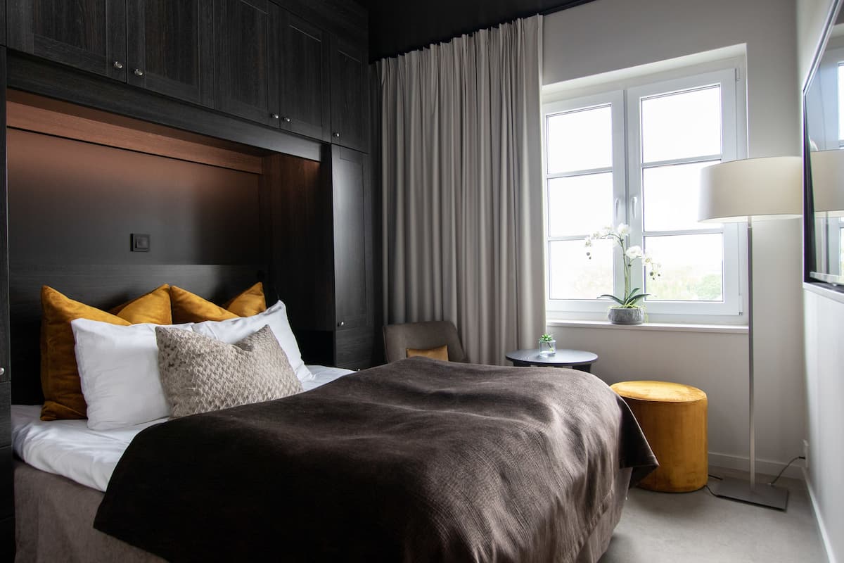 Ein Hotelzimmer mit Bett, Nachttisch, Lampe, einem Fenster mit Vorhängen und Blick ins Freie.