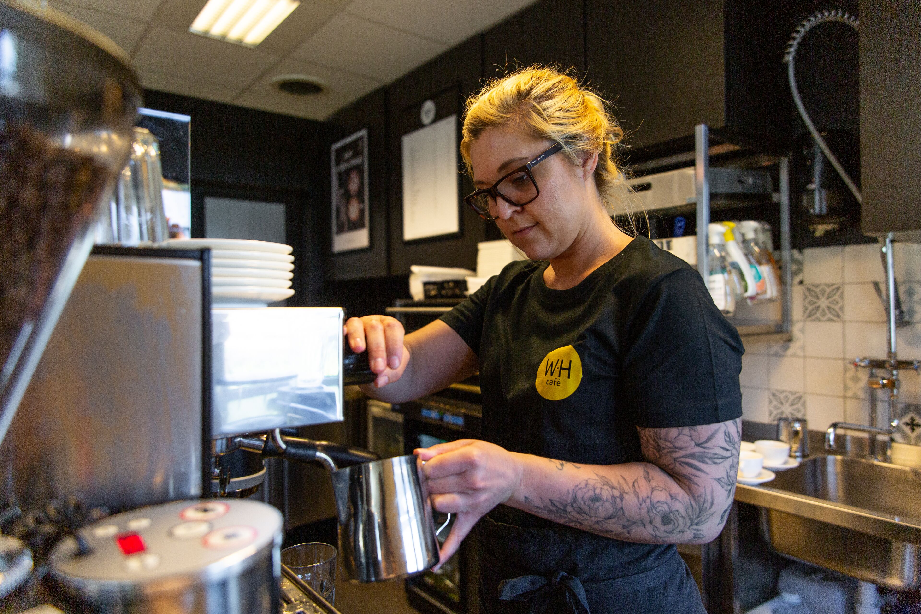 A person in a black shirt with a "WH cafe" badge, preparing a beverage in a commercial coffee machine.