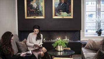 A room with two framed paintings on the wall, a fireplace, a round table with a plant, and a sofa with cushions.