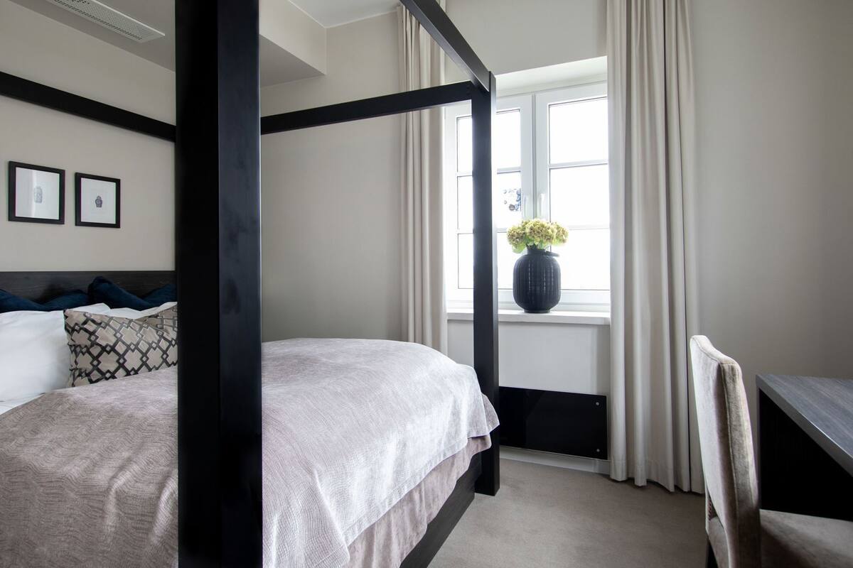 Ein Bett mit Baldachin, ausgestattet mit einer neutralen Tagesdecke, ein Fenster mit Vorhängen, eine Vase mit Blumen und ein Schreibtisch mit Stuhl.