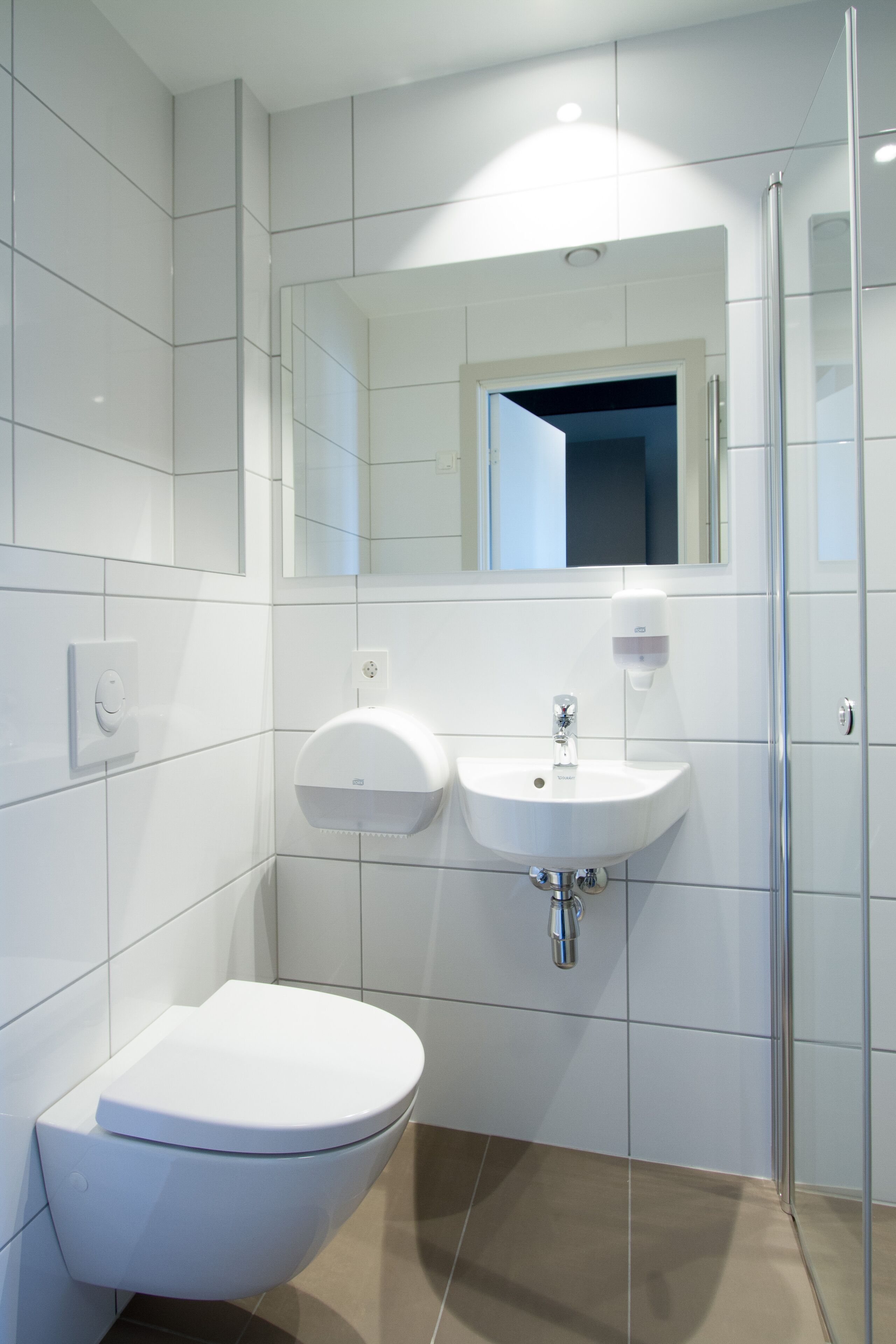 A modern bathroom with white tiles, a white toilet, a sink, and a glass shower enclosure.