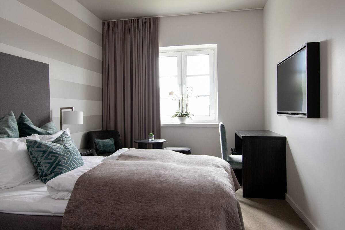 Ein Hotelzimmer mit einem Bett, einem Fernseher, einem Fenster mit Vorhängen, einem kleinen Tisch mit einer Pflanze und einem Sessel.
