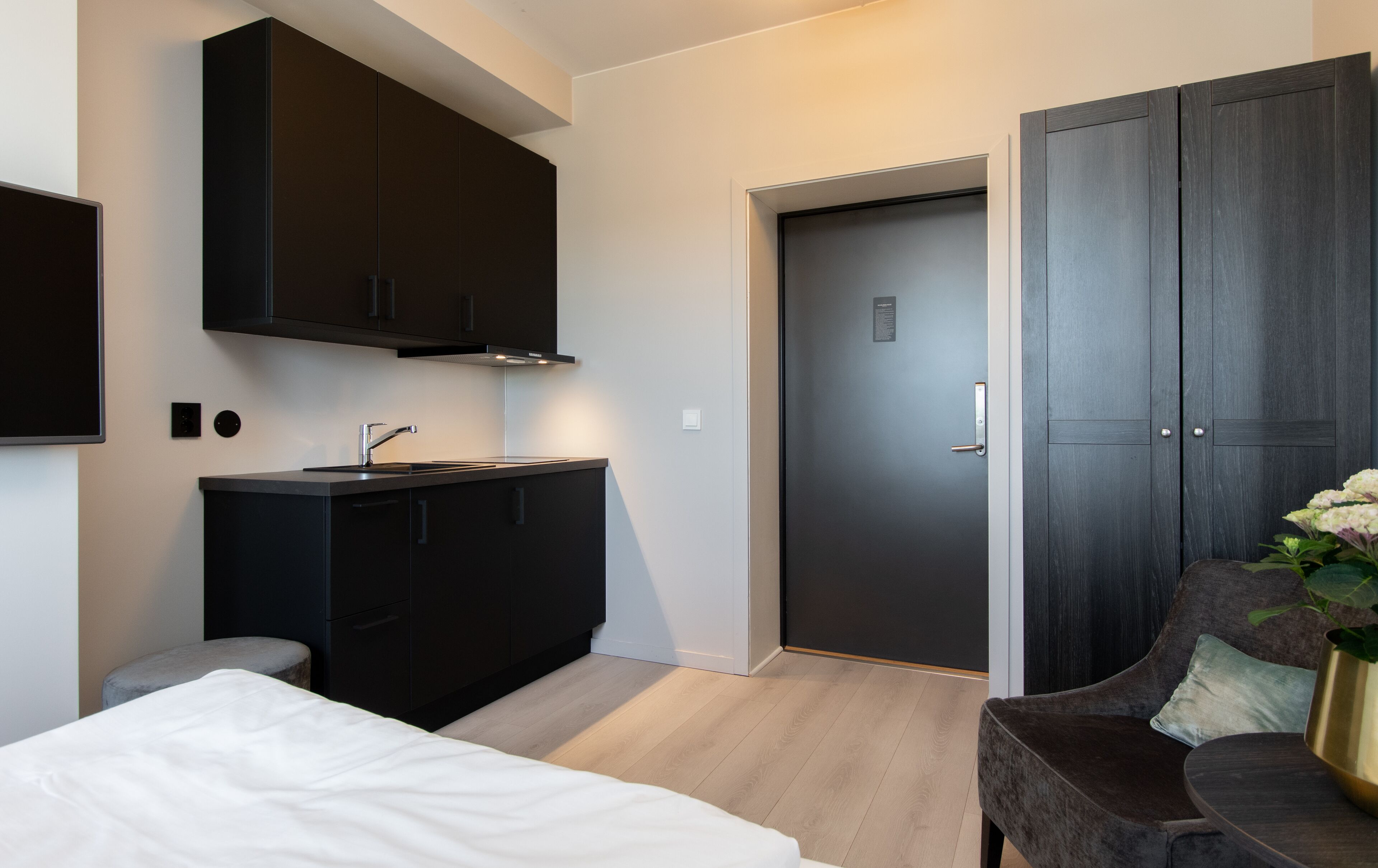 A modern bedroom with a bed, a dark wooden wardrobe, a built-in desk with a sink, and a door leading to another room.