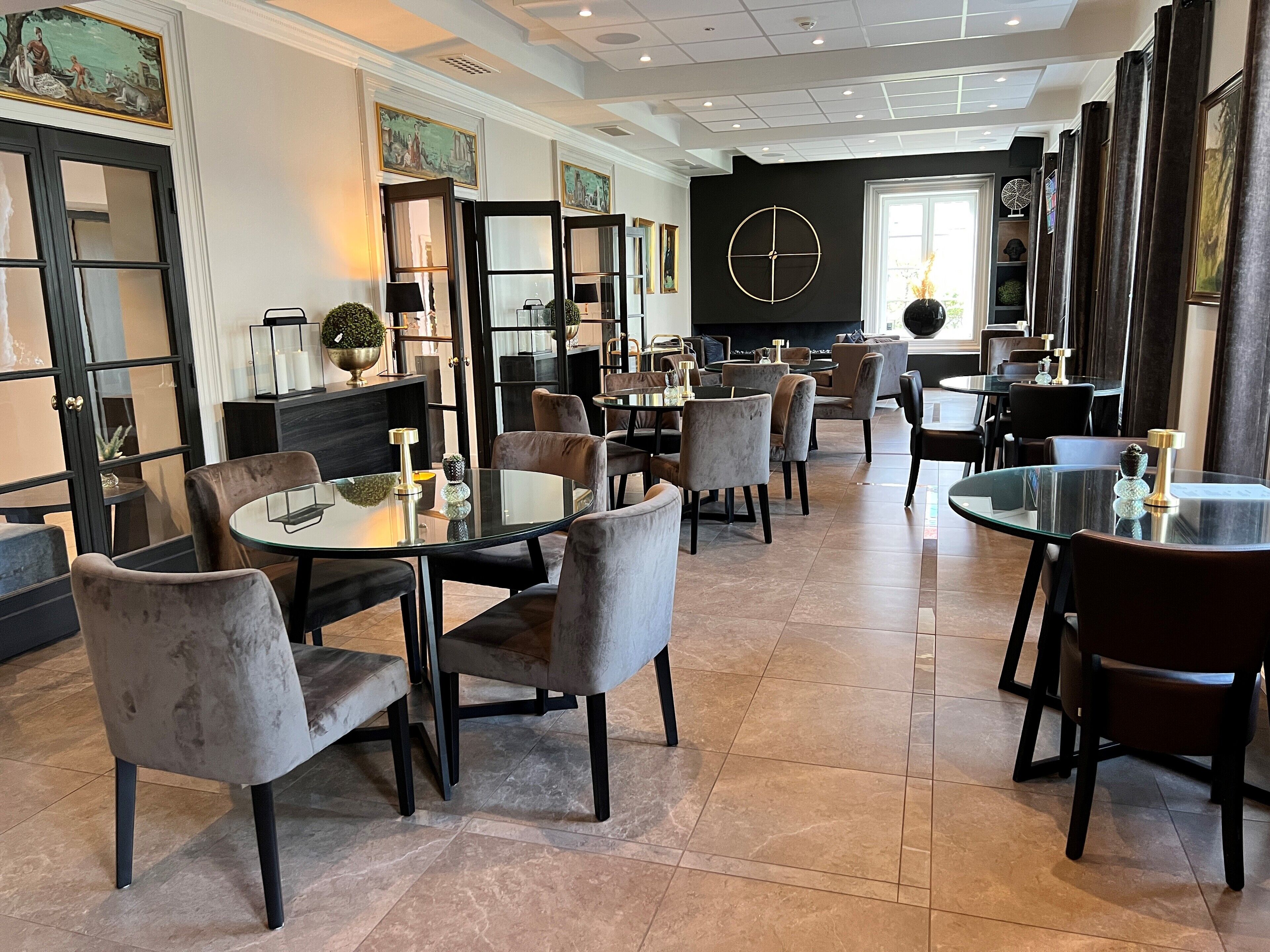 A modern dining area with round tables and velvet chairs, a black wall with a clock, and artwork on the walls.