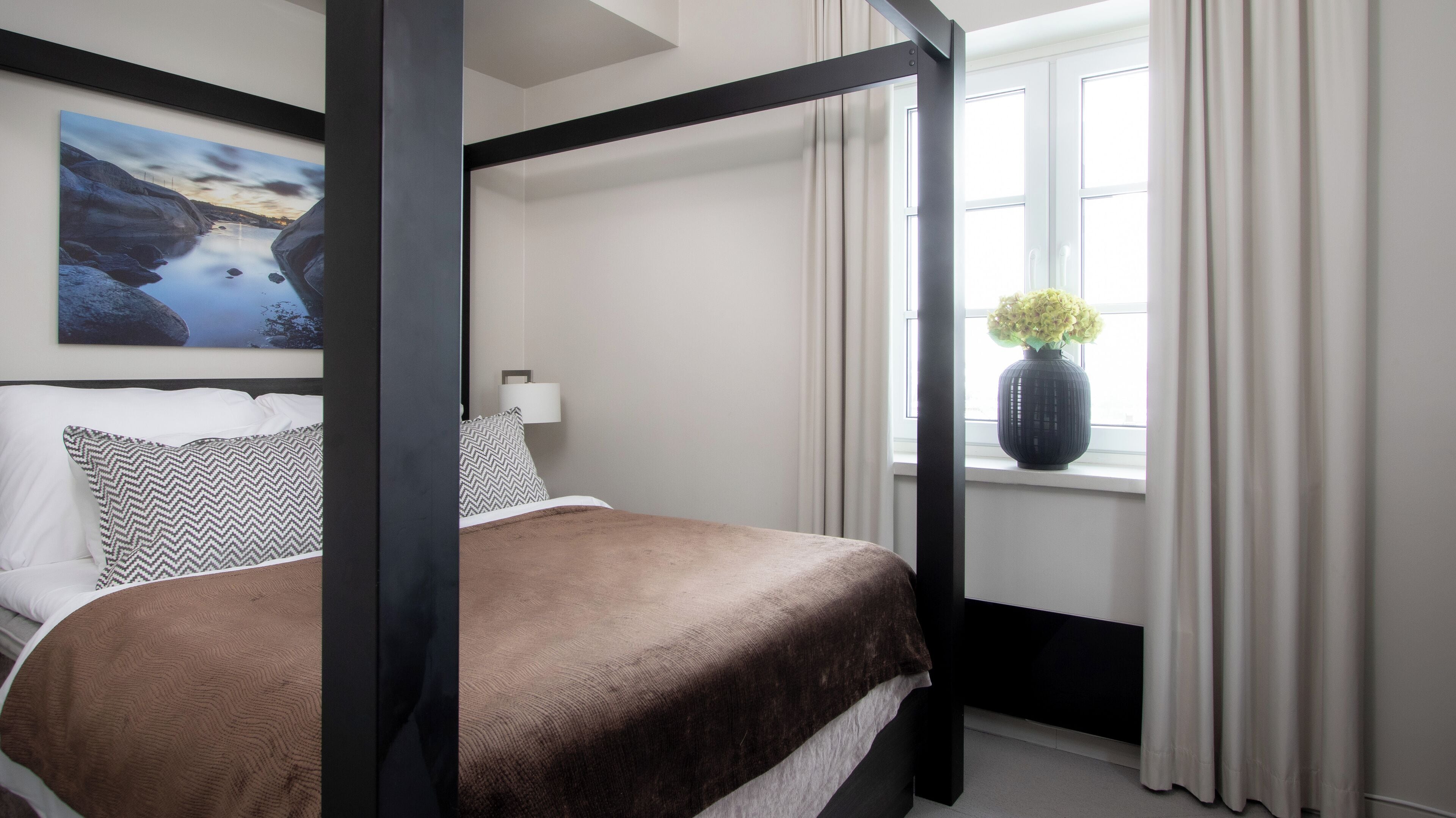 A four-poster bed with a brown bedspread, a painting of a landscape on the wall, and a vase with flowers on the windowsill.