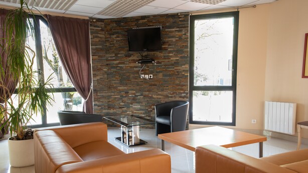 Une chambre d’hôtel moderne avec un canapé en cuir, une table basse en verre, un téléviseur à écran plat fixé à un mur en briques et de grandes fenêtres avec des rideaux.