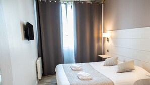 Une chambre d’hôtel avec deux lits, un téléviseur fixé au mur, une table de chevet avec une lampe et une fenêtre avec des rideaux.