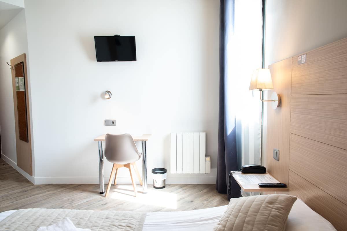 Habitación de hotel con cama, escritorio con silla, televisor montado en la pared, lámpara y radiador.