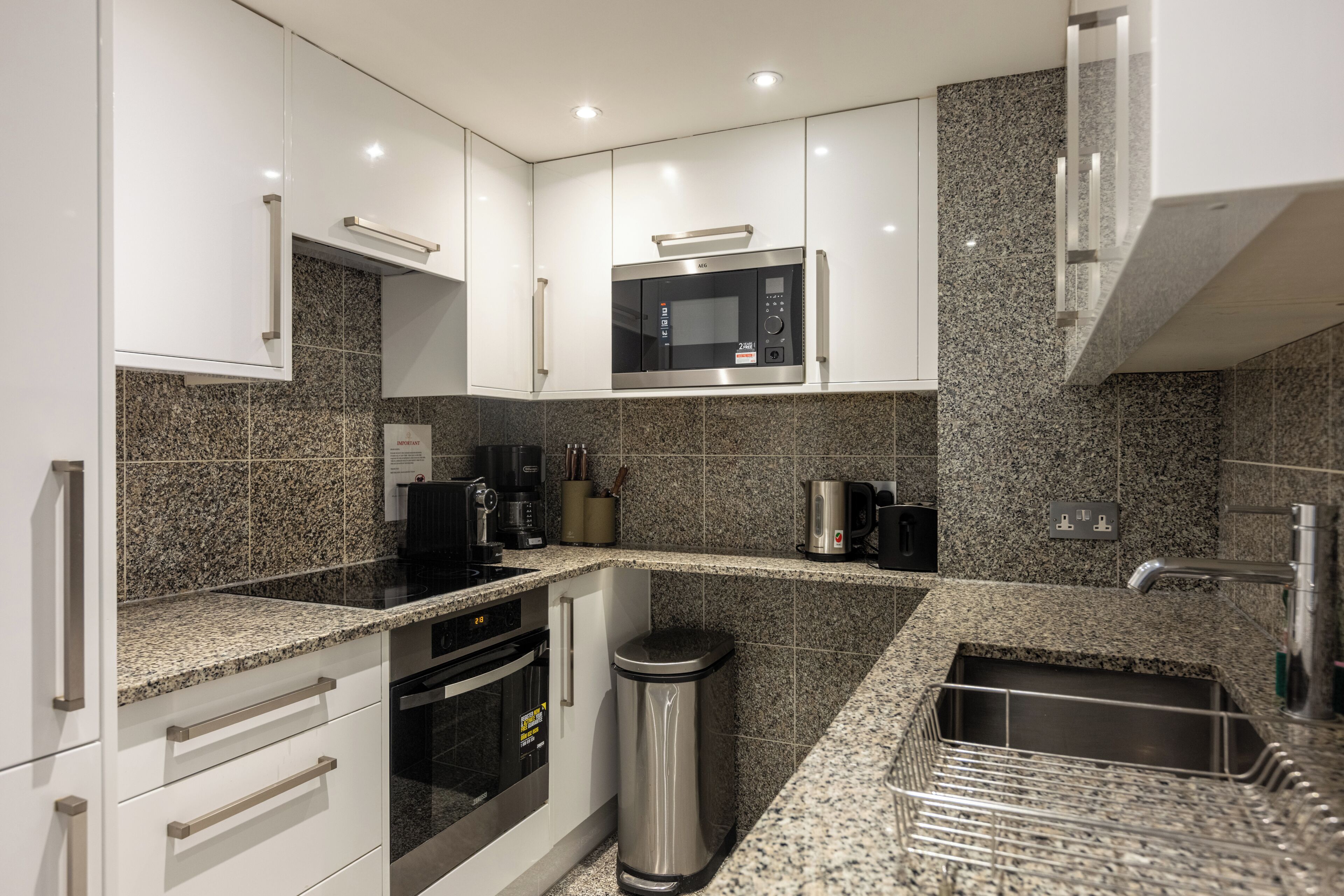 A modern kitchen with white cabinets, granite countertops, a stainless steel trash can, and a built-in microwave.