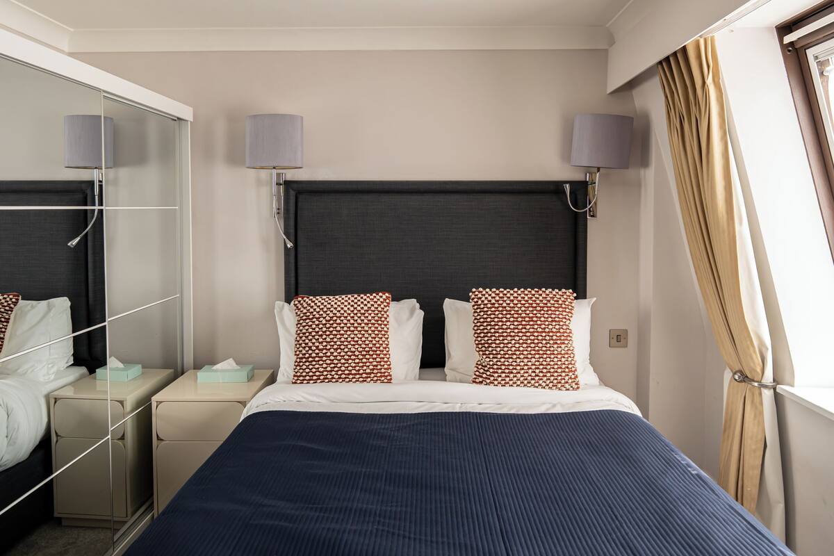 A hotel room with a large bed, a wardrobe, a mirror, a bedside table, and a window with curtains.