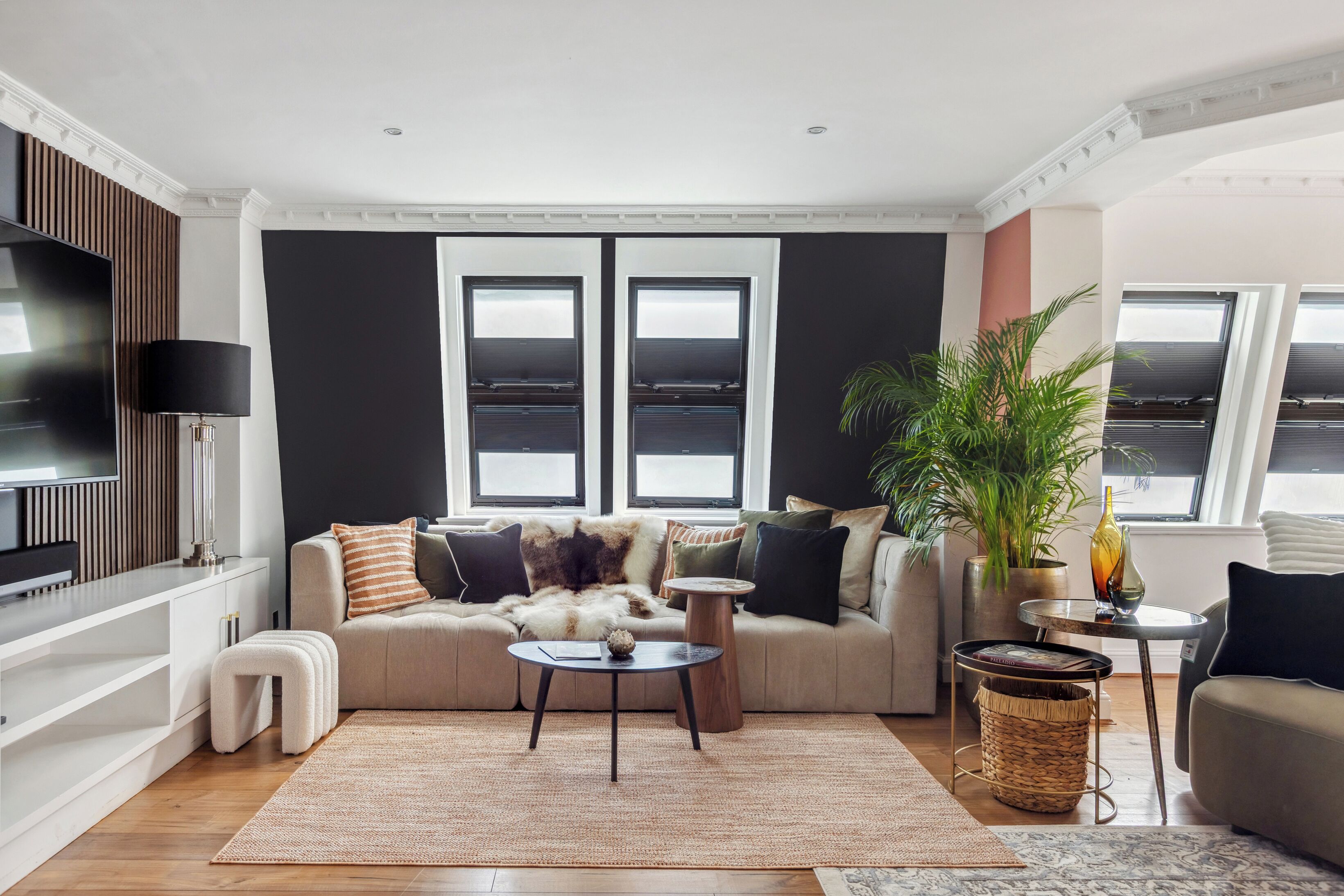 Un salon moderne avec un canapé, une table basse et un téléviseur. Il y a deux fenêtres avec des stores, une plante et une lampe.