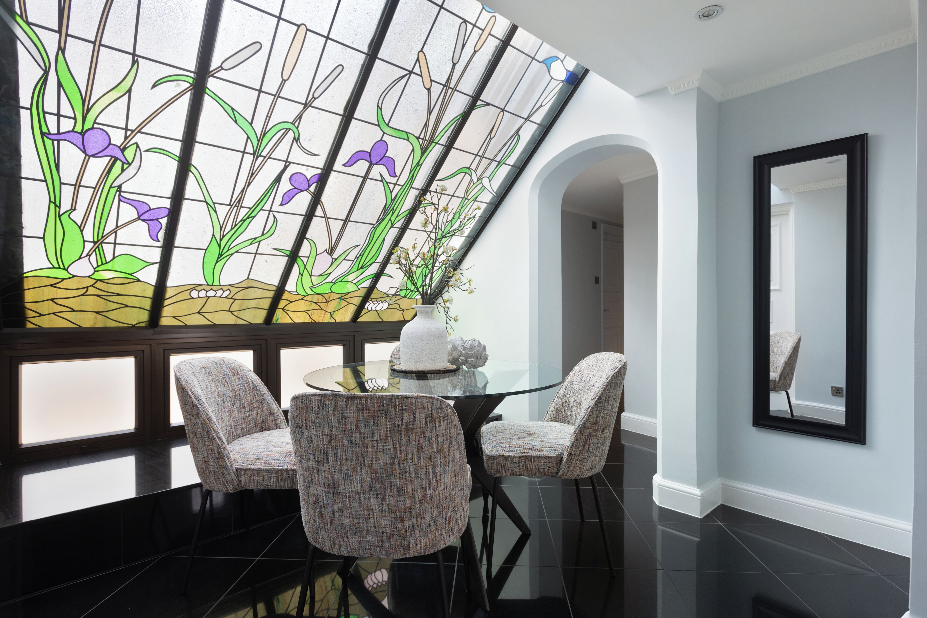 A dining area with a glass table, two upholstered chairs, a vase with flowers, and a stained glass window with plant designs.