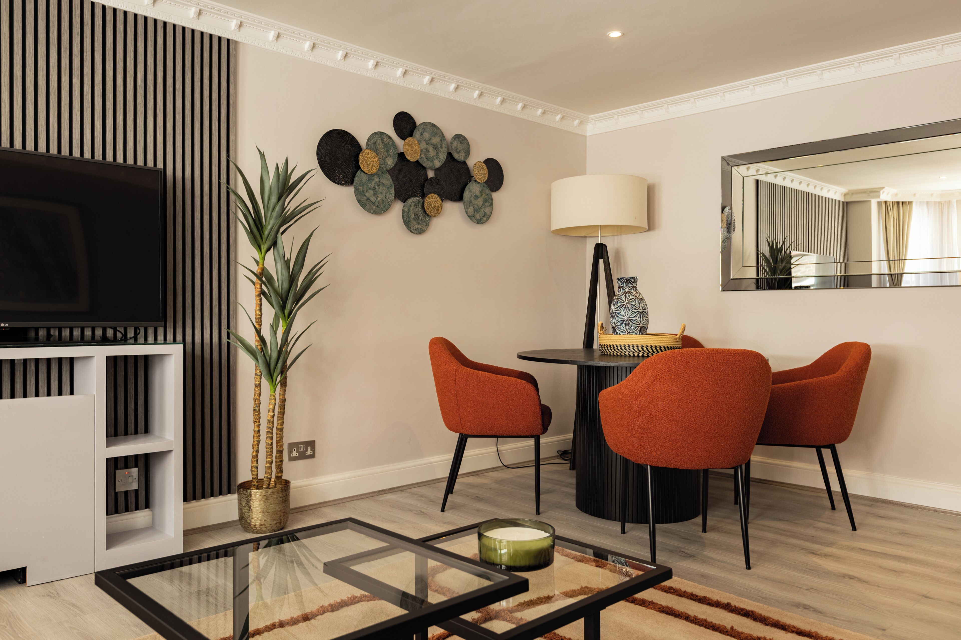 Una sala de estar moderna con televisor, mesa de comedor con sillas, lámpara y un adorno de pared.