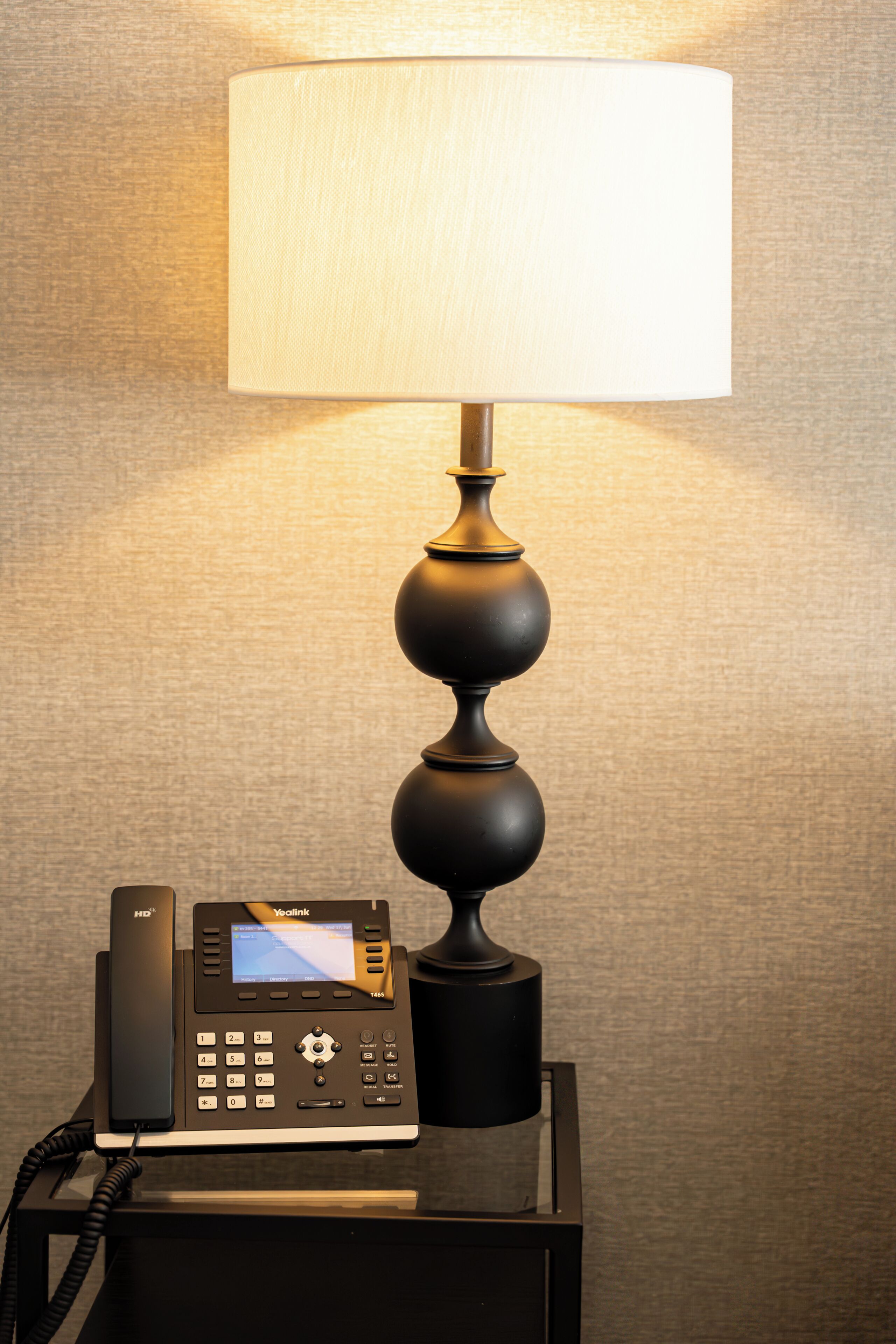 A hotel room with a desk lamp, a telephone, and a wall-mounted screen.