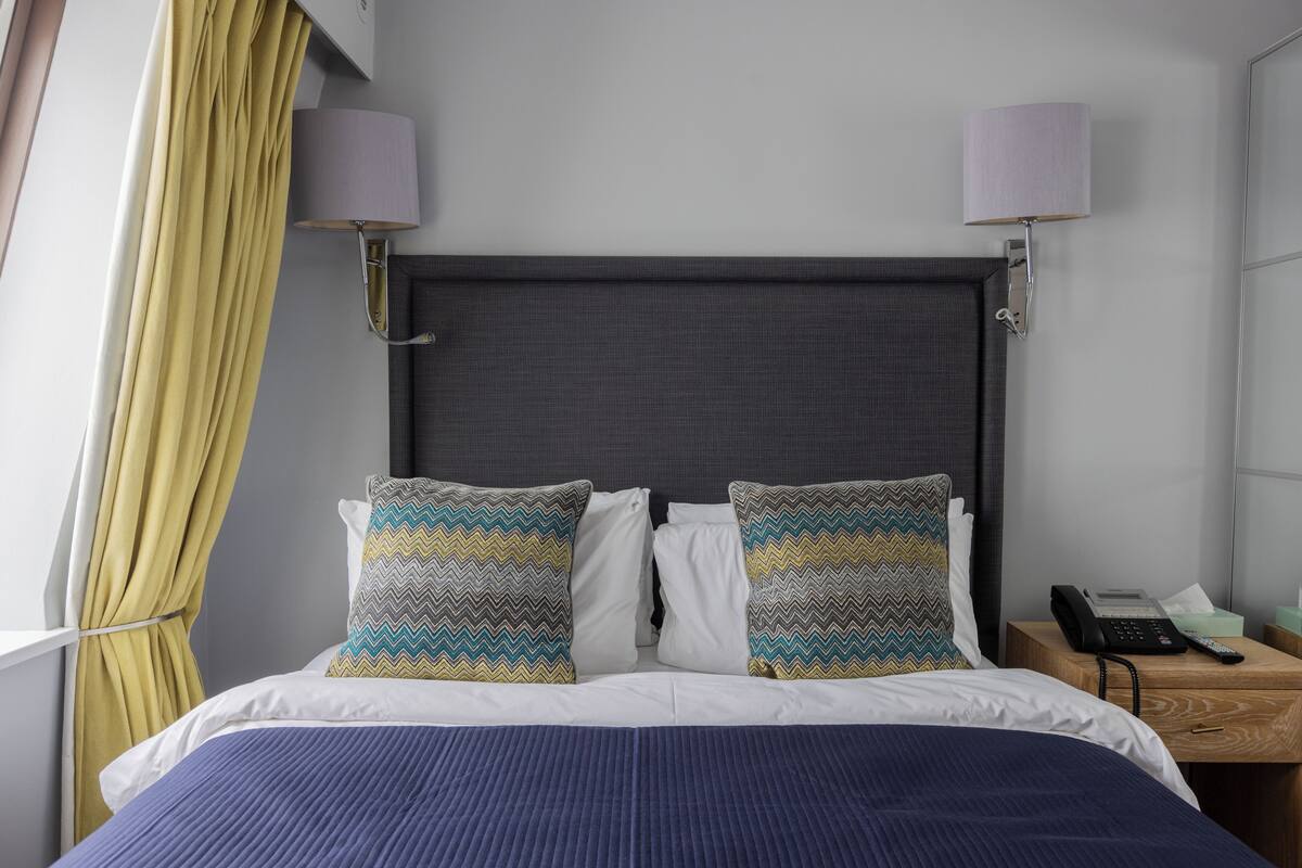 A hotel room with a bed, two wall-mounted lamps, a bedside telephone, and a window with curtains.