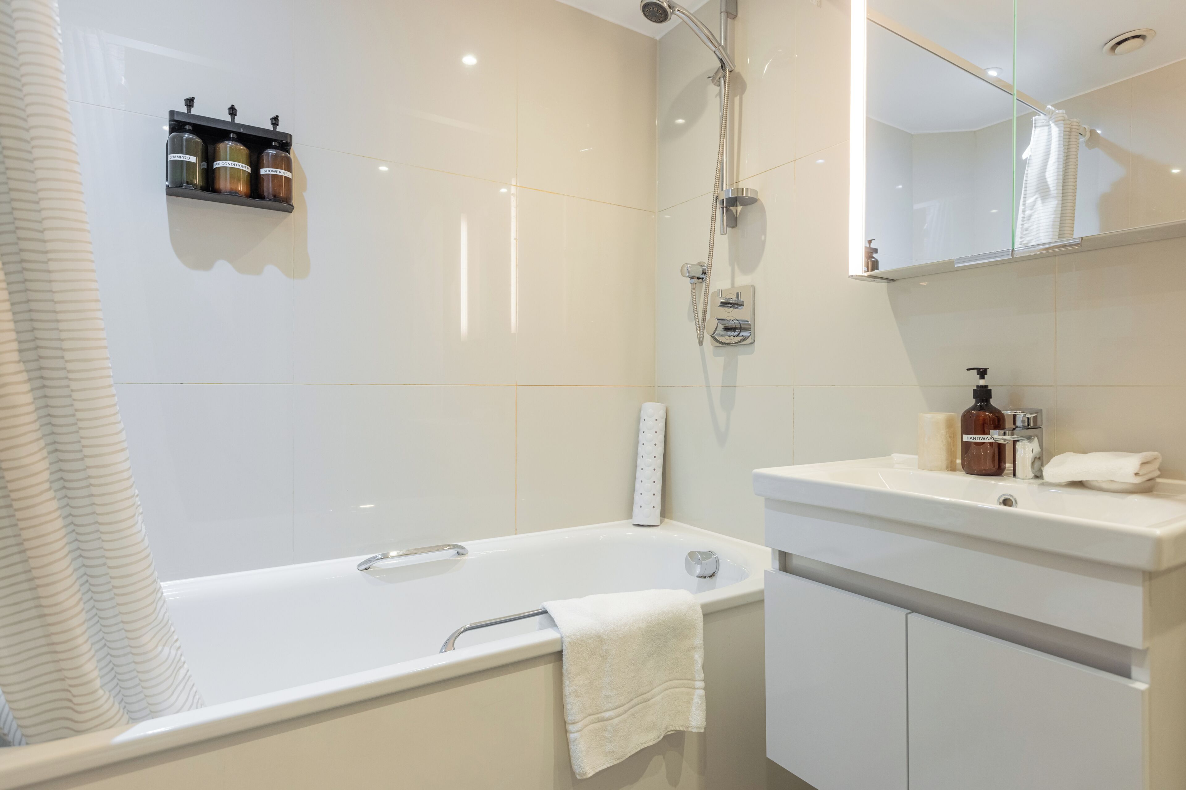 A modern bathroom with a bathtub, a shower, a vanity with a mirror, and a shelf with toiletries.