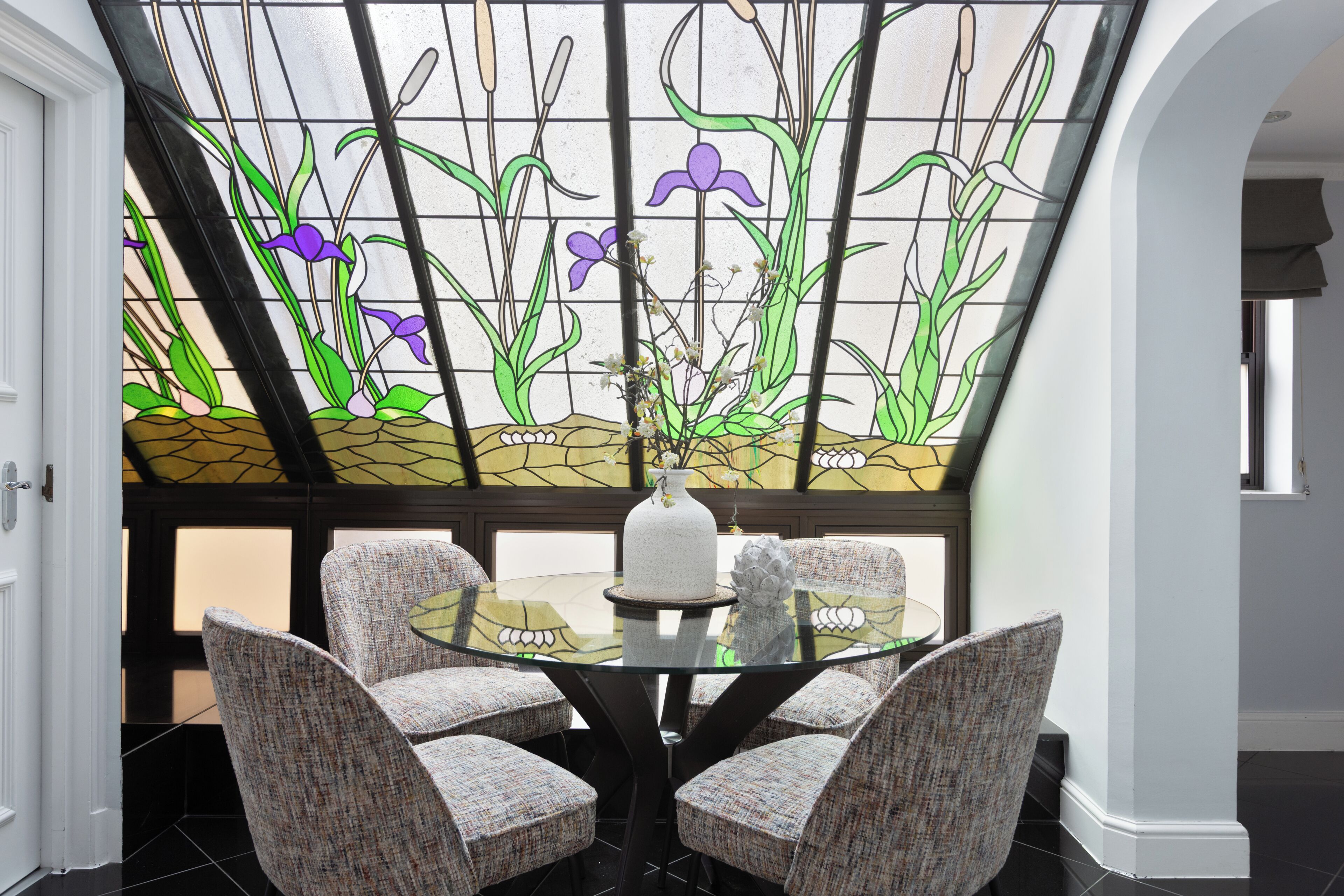A dining area with a glass table surrounded by four chairs, a vase with flowers, and a stained glass window above.