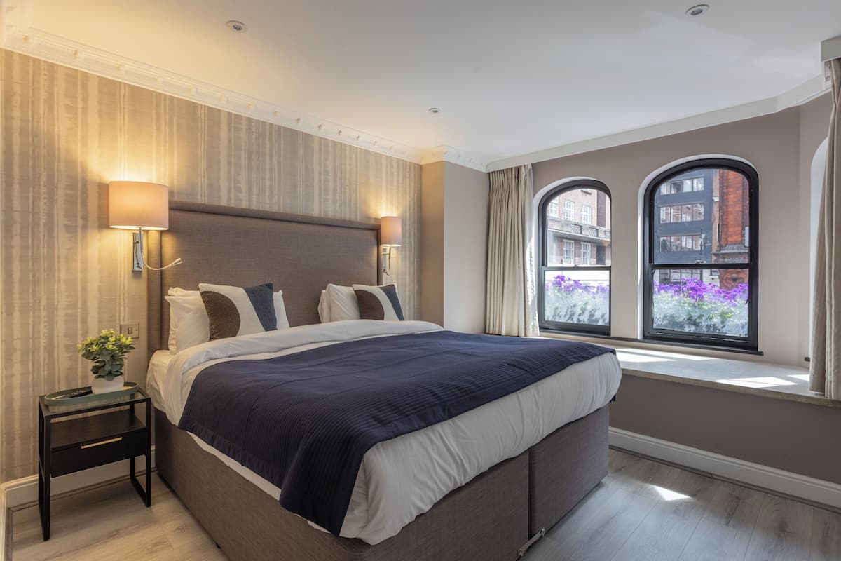 A modern hotel room with a large bed, two bedside lamps, a nightstand, and a view of buildings through two windows.