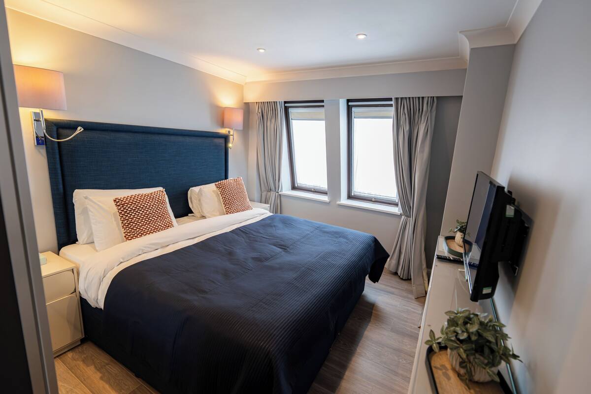 A modern hotel room with a large bed, bedside tables, a flat-screen TV, and a window with curtains.