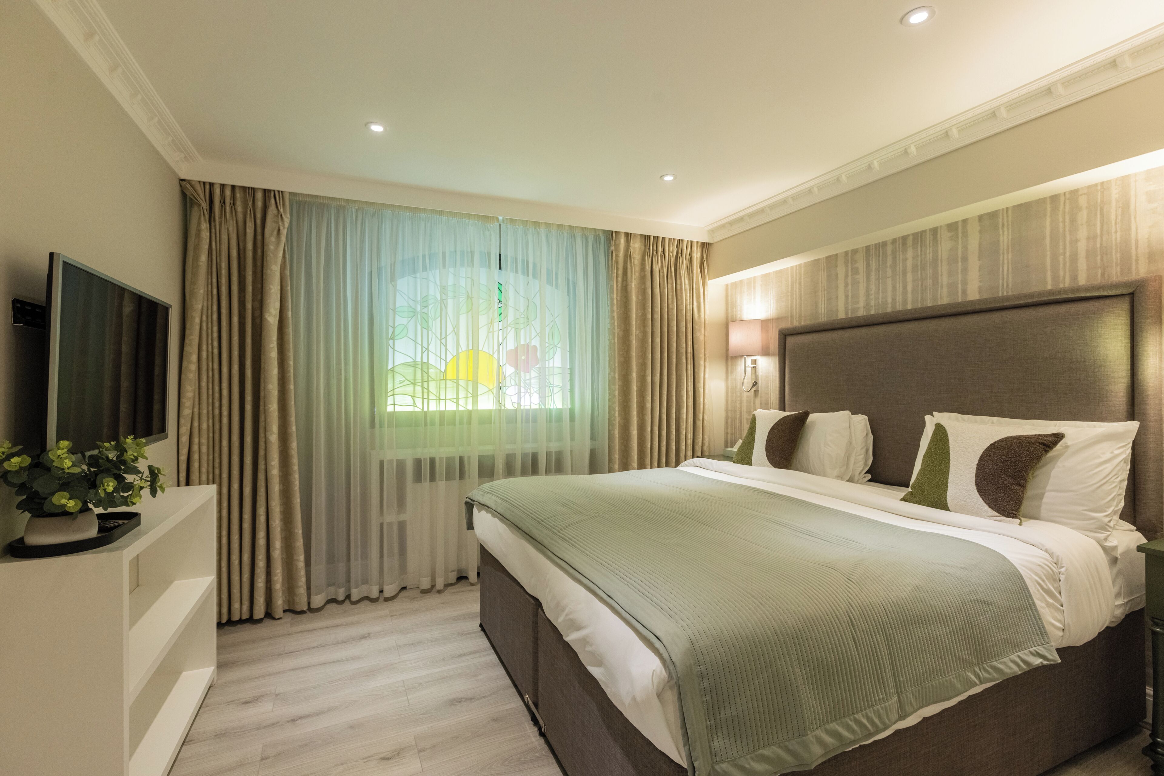 A hotel room with a large bed, a flat-screen TV, a potted plant, and a window with curtains.